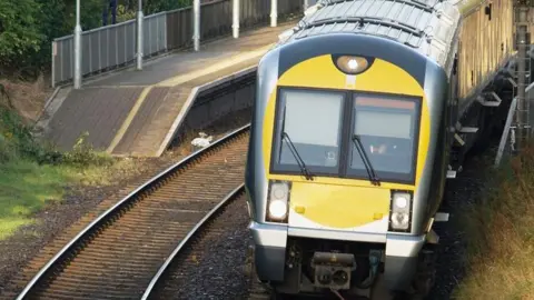 A Translink train in green, silver and yellow livery, travels along a track towards the camera