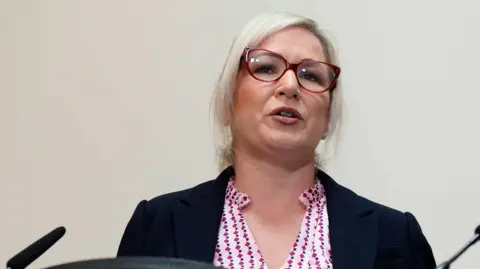 Michelle O'Neill mid speech with red glasses and pink shirt and navy blazer on a media platform