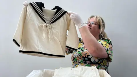 A woman wearing glasses and a floral dress holds up a children's white sailor suit top. It has navy blue trimmings, a v-neck and a bow tied across the chest.