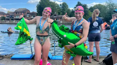 Georgia and Melissa Laurie hold green foil crocodile balloons, wearing swimming costumes and pink swim hats, smiling and have their thumbs up having just completed the swim