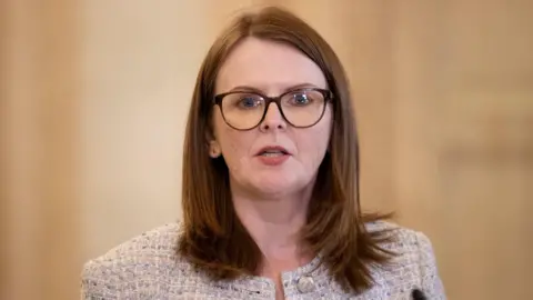 a woman with shoulder length brown hair is talking to someone off camera. She's wearing dark rimmed glasses and a light, tweed-style jacket.