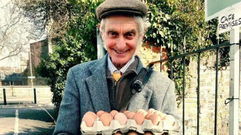 Wilf Peddle wearing a tweed cap and blazer holding a carton of eggs and smiling