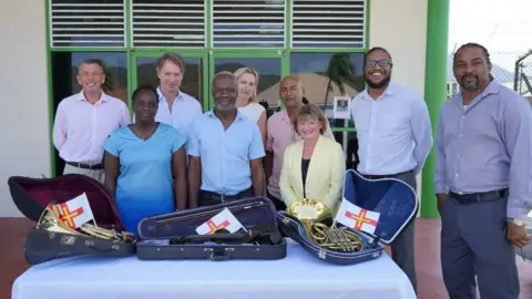 Representatives of Guernsey Music service pose with organisers of a Montserrat music summer school. they are standing as a group, smiling for the camera, standing behind a table with a white table cloth which has three opened musical instrument boxes on it. There are three small Guernsey flags in the boxes.