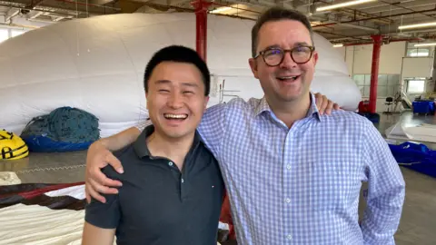 Dennis Hu and Ben Hardy with their arms round each other's shoulders, smiling, while a white balloon is partly inflated behind them.