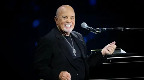 Billy Joel on stage smiling, he is imitating holding a guitar, the piano is to his right and a mic is in front of him