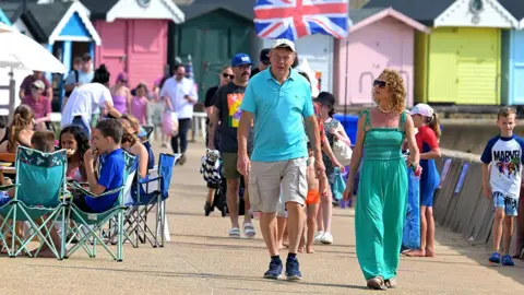 A couple walk down the promenade in summery clothing, with one wearing a sun hat and the other sunglasses, with beach huts and the union flag in the background, in Walton on Sea on 12 August