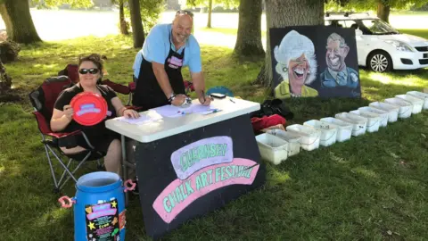 A man and a woman smiling behind a desk which says GUERNSEY CHALK ART FESTIVAL next to a line of white bucket full of chalk, a caricature of the King and Queen, in a field of trees and a white car on the right, sunny day