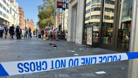 Blue and white police tape in front of the scene of the stabbing with bits of white paper and cloth on the ground and a crowd of people