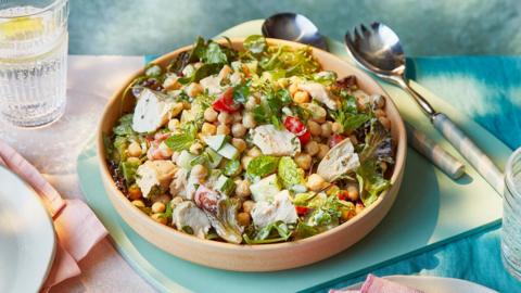 A bowl of colourful salad including chickpeas and chicken next to serving spoons and a glass of water with lemon it in.