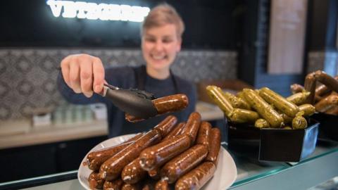 Someone smiling while holding a sausage with a pair of tongs over a counter top of several plates of sausages.