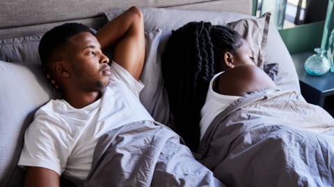 A couple in bed. The woman is facing away and is asleep while the man is awake with his hand behind his head looking over at the woman.