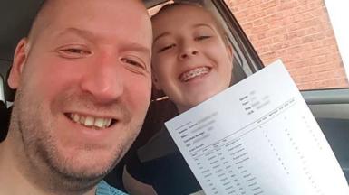 Anna smiles as she holds up her A-level results, printed on a white sheet of paper. She is sat next to her dad, Les Sutton, in a car